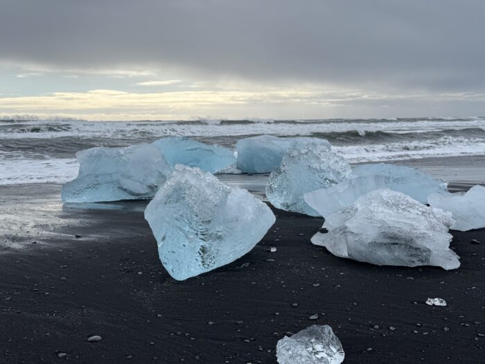 Ice chunks on black sand beach, Iceland, with ocean waves and overcast sky in background.
