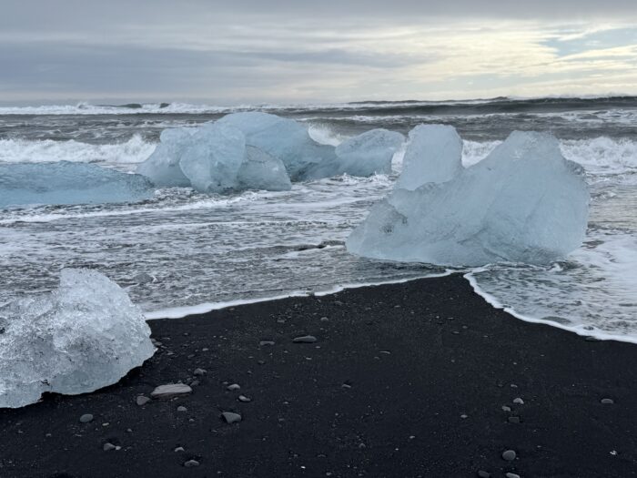 Icebergs on black sand beach in Iceland with waves. Cloudy sky, blue ice, black sand.