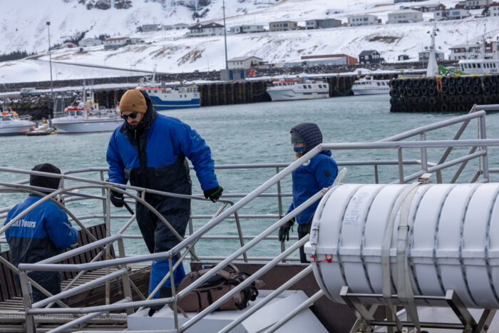 People in blue jackets on a boat deck in a harbor with snow-covered hills.
