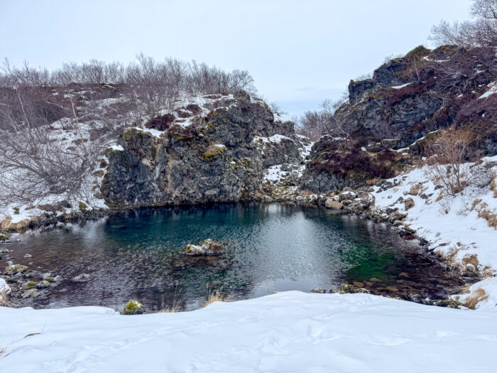 Turquoise hot spring surrounded by snow-covered rocks and bare trees in Iceland.